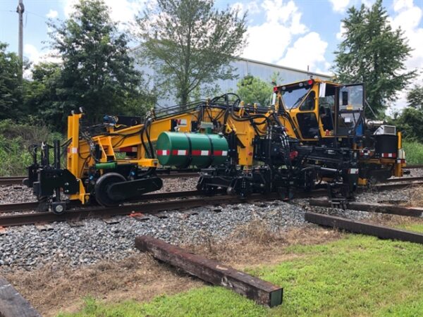 A Nordco TPX Tie Exchanger actively removing ties from a rail and placing them in the ballast